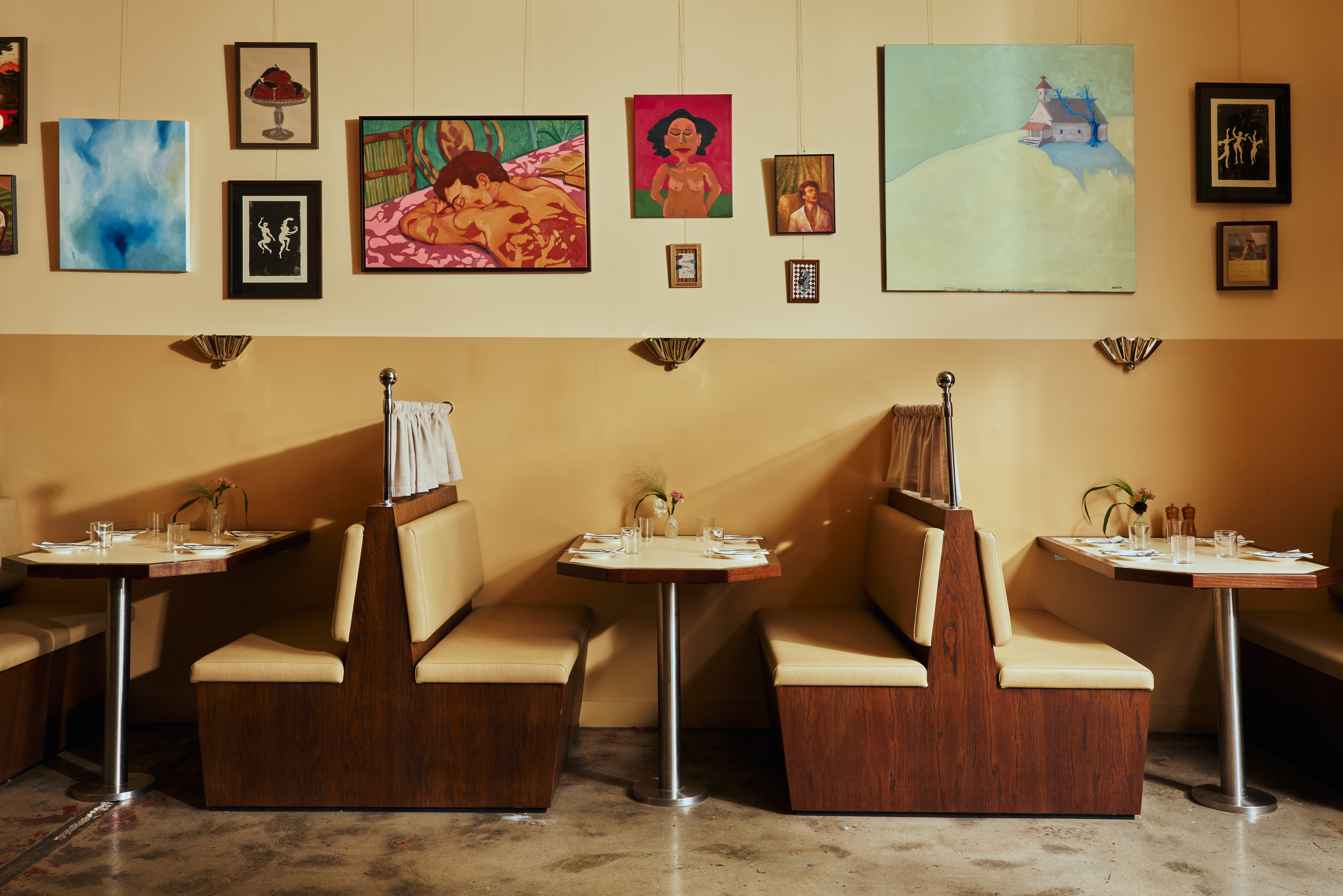 A restaurant banquette booth made of pale, camel-tinted leather, dark wood, and a table crafted with the same materials except for its chrome legs sits in a yellow-painted restaurant with stone floors, topped by two espresso cups, an open book, salt and pepper grinders in wood, a tiny flower vase, and a chrome table lamp.