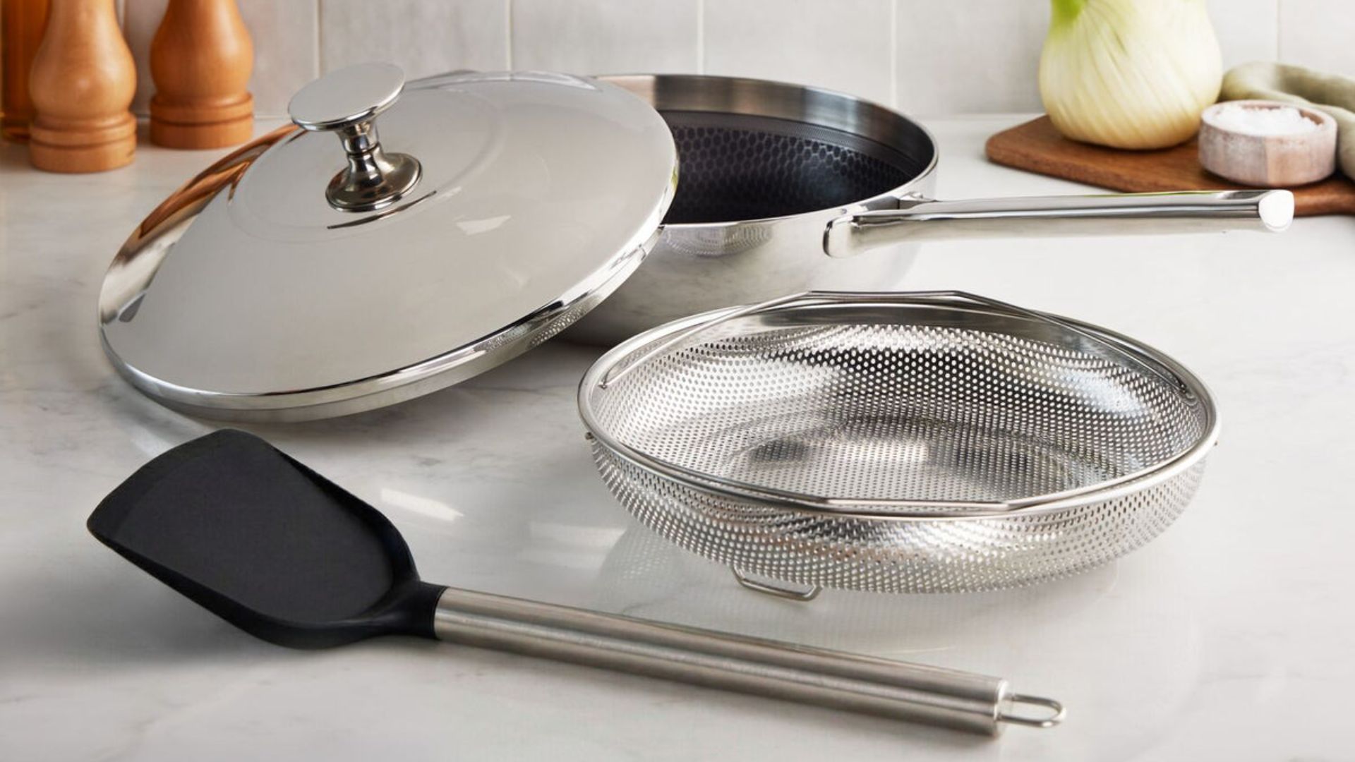 Lakeland 10-in-1 Tri-Ply Only Pan Pro on a white background on a counter with the steamer and spatula in front at the lid propped on top