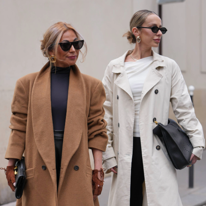 PARIS, FRANCE - OCTOBER 04: A guest (L) wears a loose low bun hairstyle with honey-blonde highlights and face-framing tendrils, black cat-eye sunglasses, large gold hoop earrings, a black leather clutch bag, a camel wool long double-breasted coat with wide lapels and black buttons over a black high-neck knit top and black leather trousers, brown suede gloves / mittens, black patent leather pointed-toe pumps shoes, A guest (R) wears a slicked-back low bun hairstyle with medium-length light brown hair and a center part, black oval sunglasses, chunky gold hoop earrings and a thin gold chain necklace, a black pebbled leather shoulder oversized Evelyne handbag from Hermes with gold hardware, a long beige cotton trench coat with double-breasted black buttons worn open over a white draped asymmetric top and black wide-leg trousers, black patent leather pointed-toe pumps shoes with stiletto heels, outside Hermes, during Paris Fashion Week - Womenswear Spring Summer 2026, on October 04, 2025 in Paris, France (Photo by Edward Berthelot/Getty Images)