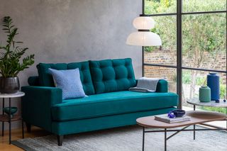 A small teal couch for a small living room, in front of a concrete wall and large crittall windows.