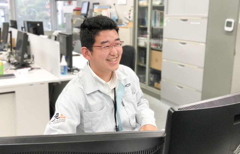 Satoshi Araki smiling while working on a computer at his office desk.