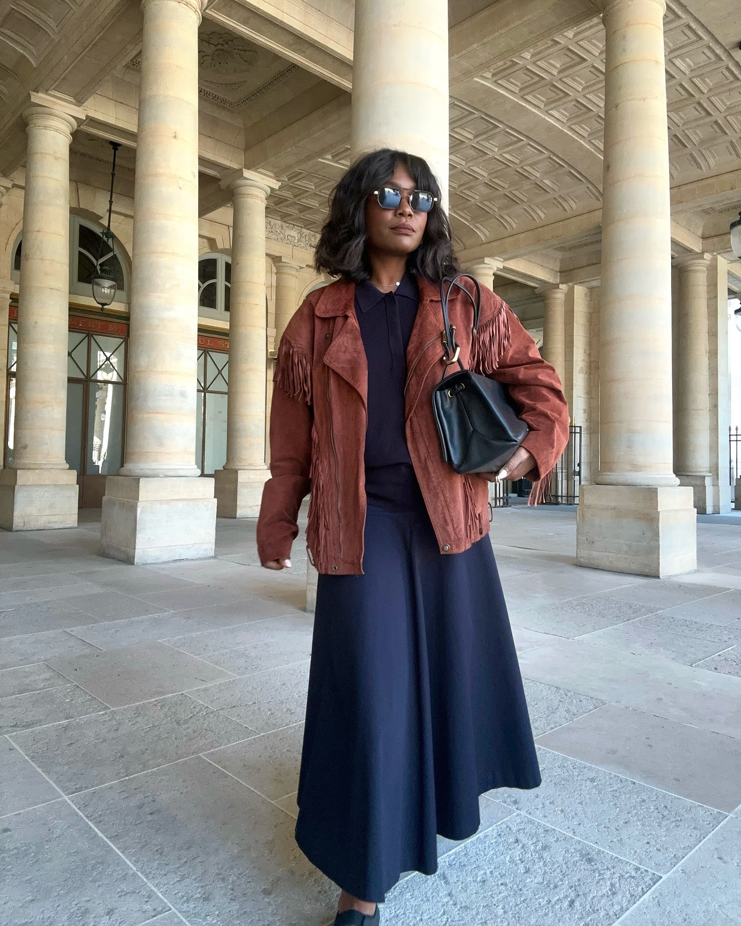 Parisian woman wears a brown suede fringed jacket with a navy polo shirt, full skirt and sunglasses.