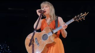 Taylor Swift wearing orange dress and holding a guitar during surprise song section of her Eras Tour. 