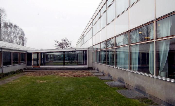 A grass and sand school courtyard flanked by two-storey building to the right and single-storey buildings on the other sides of the courtyard.