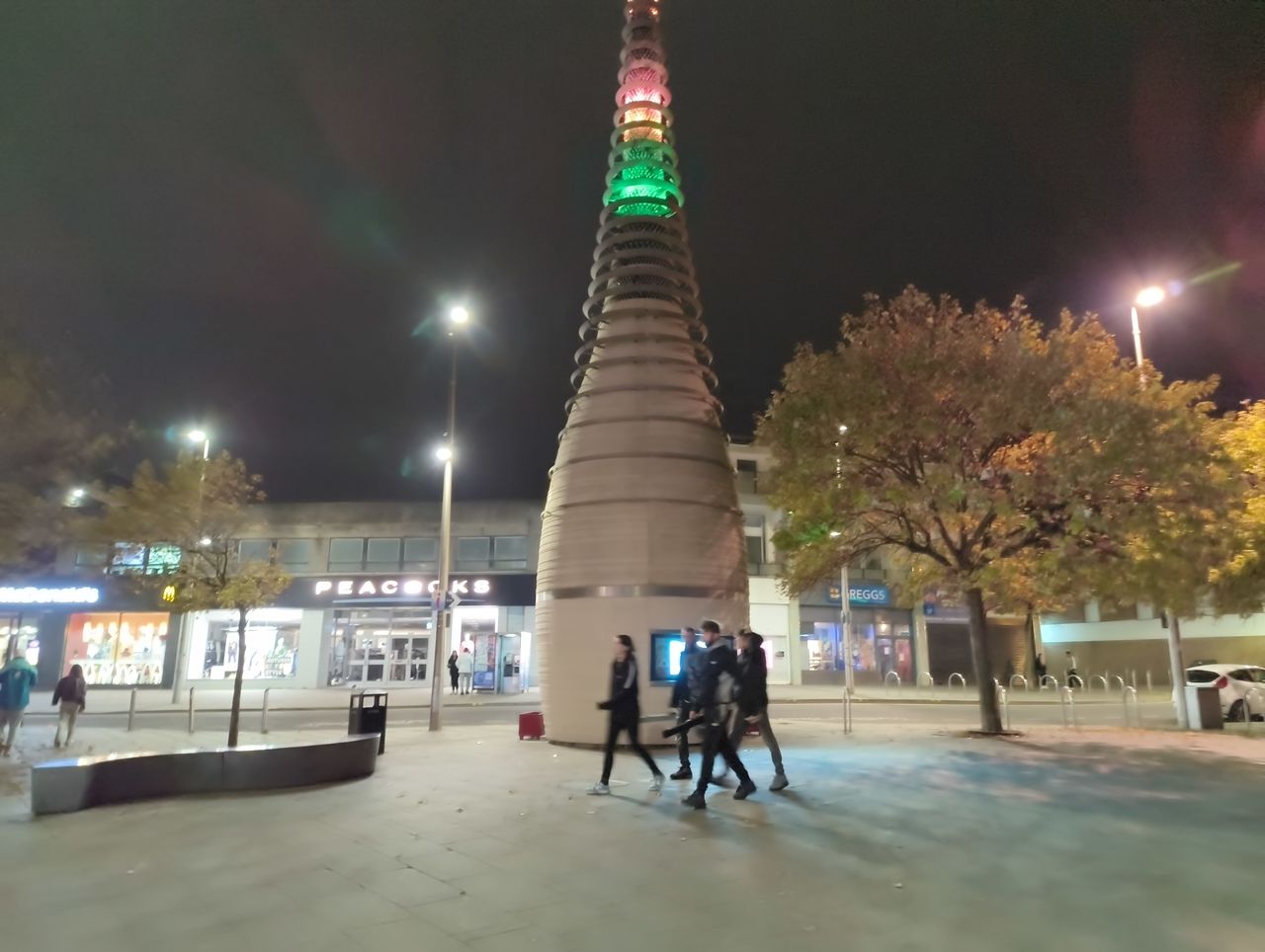 A view of a public square at night, dominated by a large, cone-shaped tower structure illuminated with colored lights at the top, with people walking below and shops in the background.