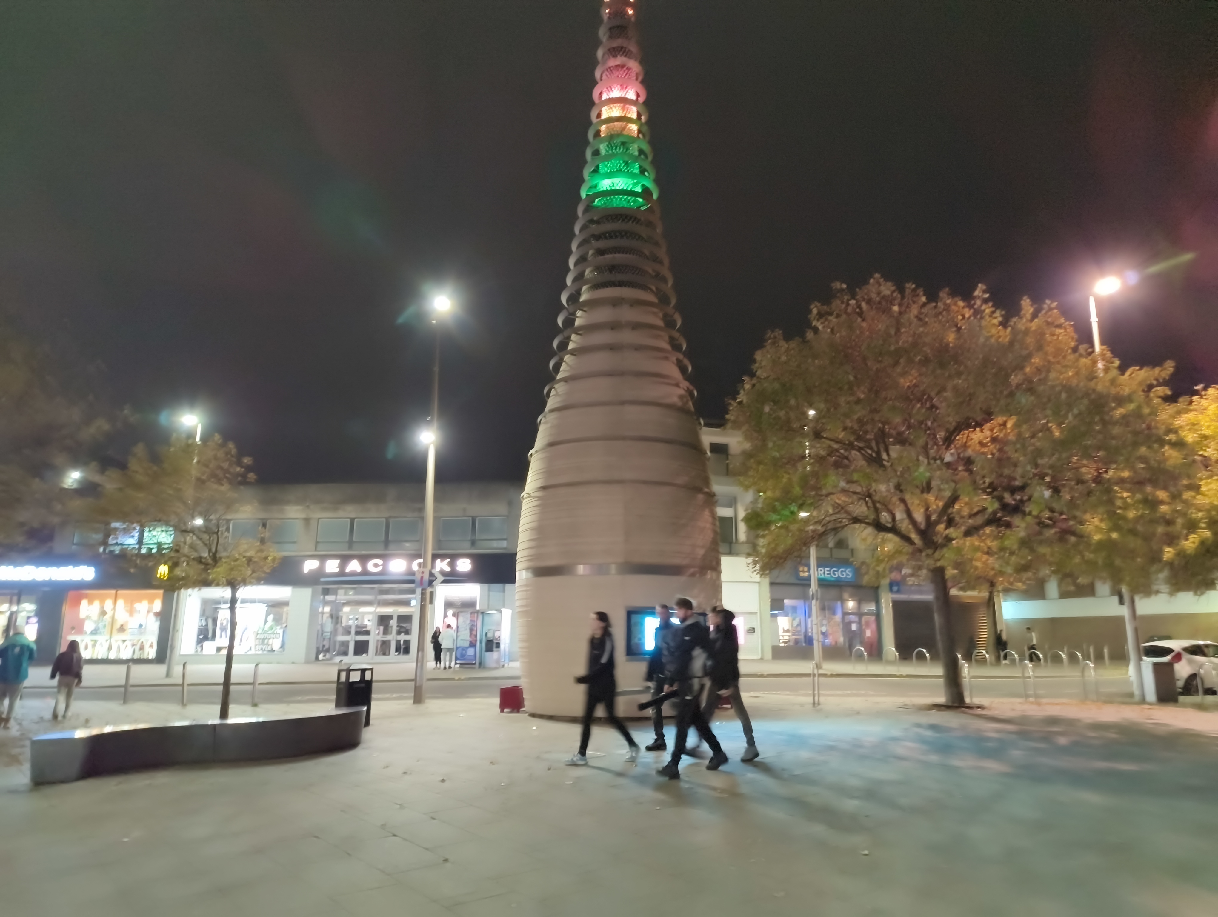 A view of a public square at night, dominated by a large, cone-shaped tower structure illuminated with colored lights at the top, with people walking below and shops in the background.