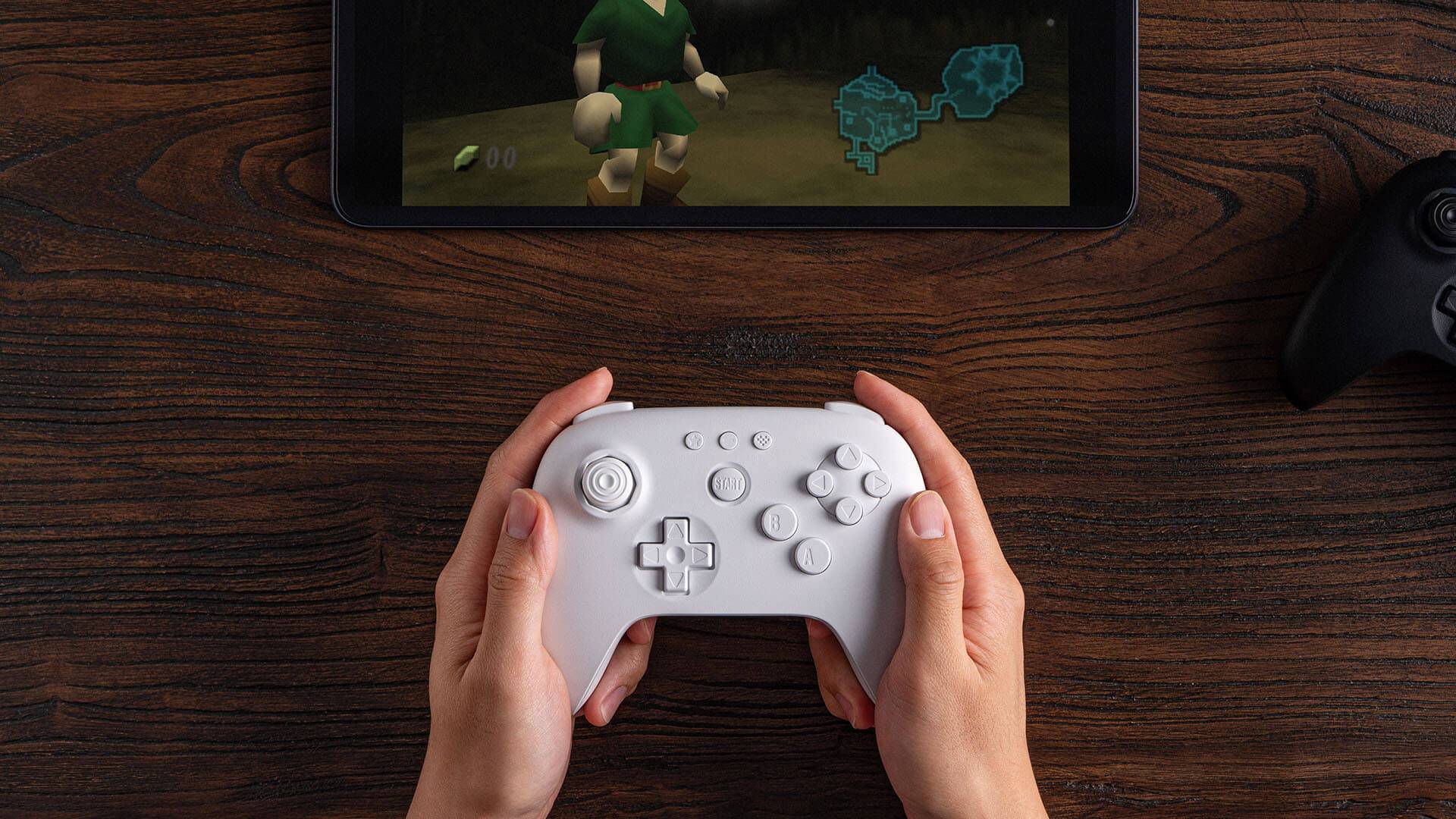 Hands holding white 8Bitdo N64 Bluetooth controller with Zelda: Ocarina of Time gameplay on tablet in front of player.