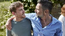 A father puts his arm around the shoulders of his adult son while they're at a party.