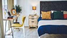 A modern Scandi-style bedroom with a desk against the window and a wooden nightstand and a blue and yellow bed