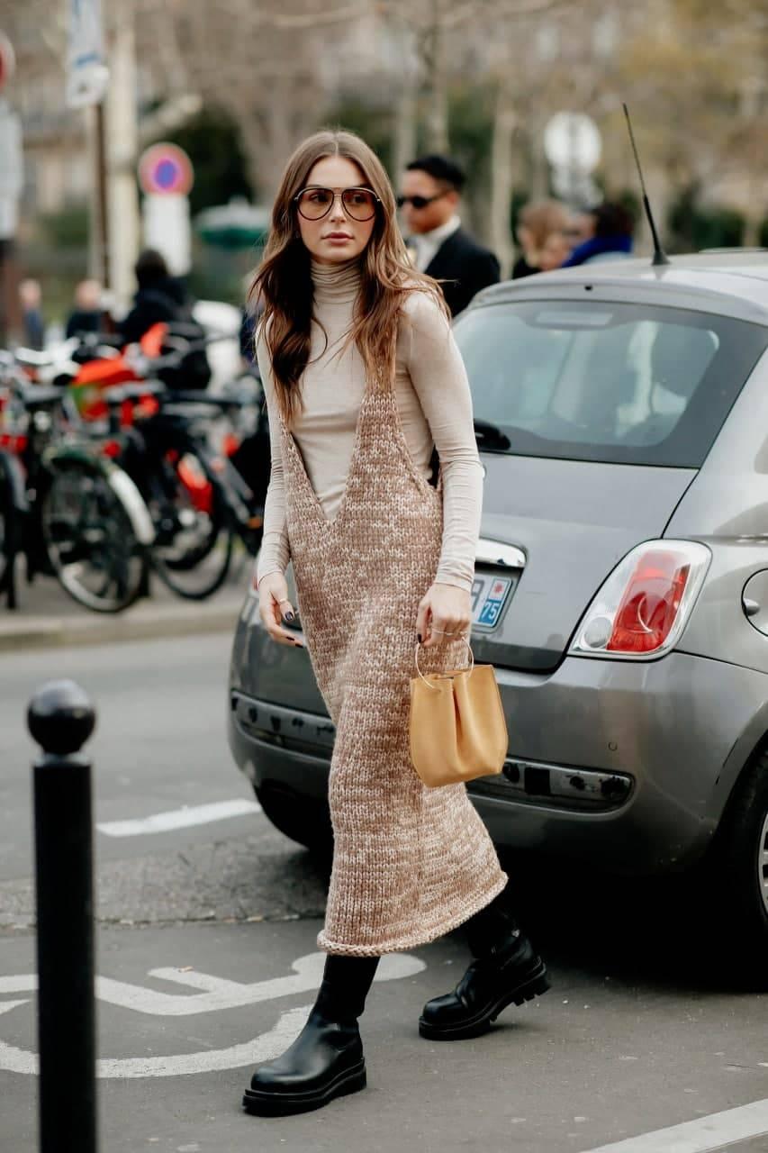A woman wears a beige turtleneck layered under a brown knit midi dress with black ankle boots and a tan handbag.