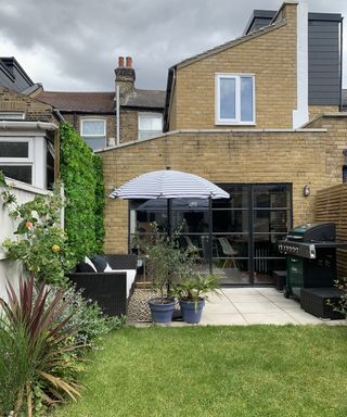 An image of a backyard with lawn laid with blue and white striped parasol umbrella, potted plants, barbecue grill in corner