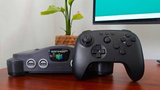 8Bitdo N64 Bluetooth Controller for Analogue 3D in black leaning against original Nintendo 64 on woodgrain desk with plant and monitor in backdrop.