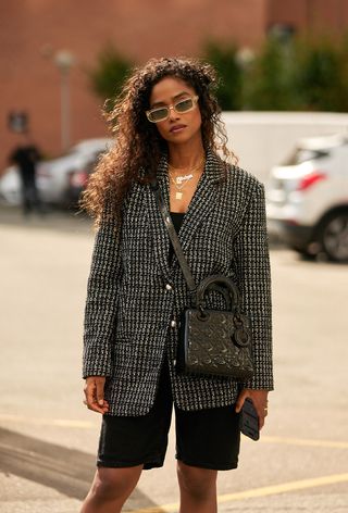 a close street style photo of a woman wearing a vintage dior bag with a black tweed blazer layered over a black tank and black shorts