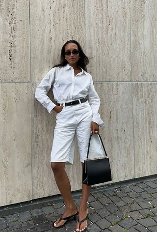 a photo showing what to wear to work during the summer with a woman wearing a white button-down shirt with white shorts and a black belt and a black bag and black thong sandals