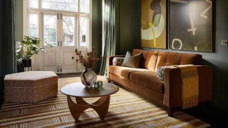 A cozy living room with dark green walls, French doors leading outside, green curtains, a burnt orange sofa with an ochre and cream modern patterned rug, a round wood coffee table, and two dark yellow and green abstract artworks on the wall. 