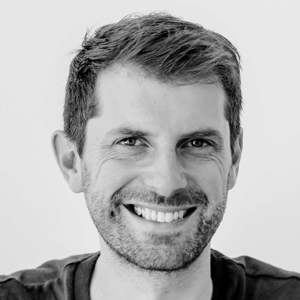 black and white headshot of male with short dark hair and facial hair, smiling and wearing dark coloured top