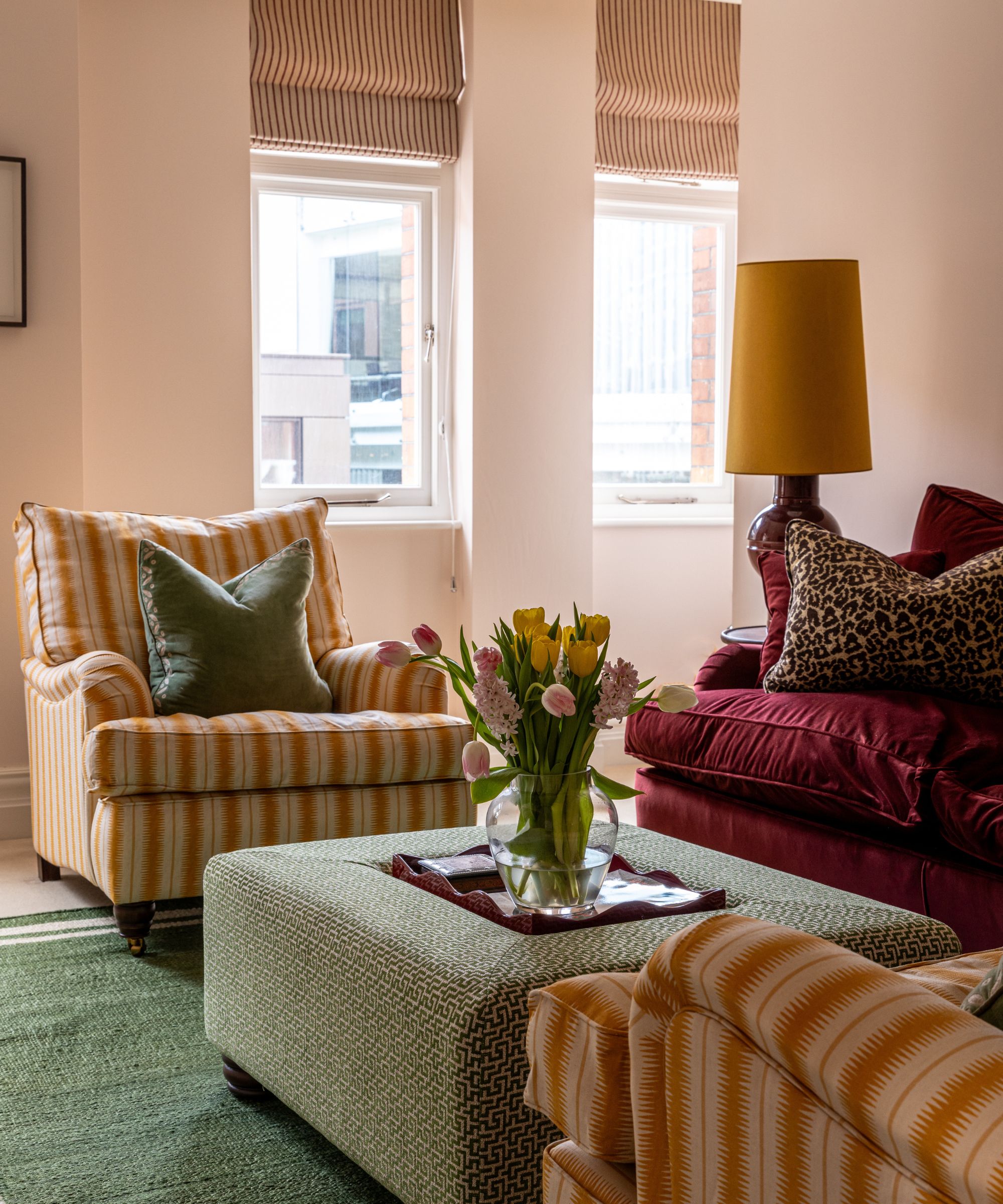 Living room with pale pink walls, striped yellow armchairs, green rug, green ottoman with lacquer tray and vase of tulips in the middle, a dark pink velvet sofa and a red lamp with a yellow shade