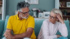 An older couple look at each other unhappily while sitting on their sofa.