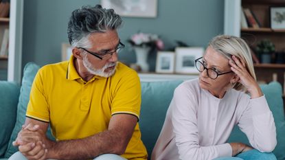 An older couple look at each other unhappily while sitting on their sofa.
