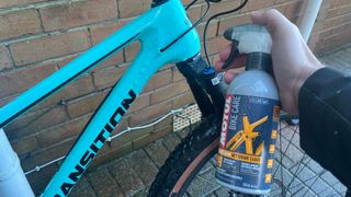 Hand holding Motul Frame Clean bottle by front of bike frame with brick wall behind