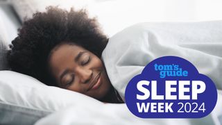 A woman with dark curly hair closes her eyes and smiles as she lays her head on a white pillow and pulls a white duvet up to her chin. A Tom's Guide Sleep Week 2024 logo is placed over the top of the image.