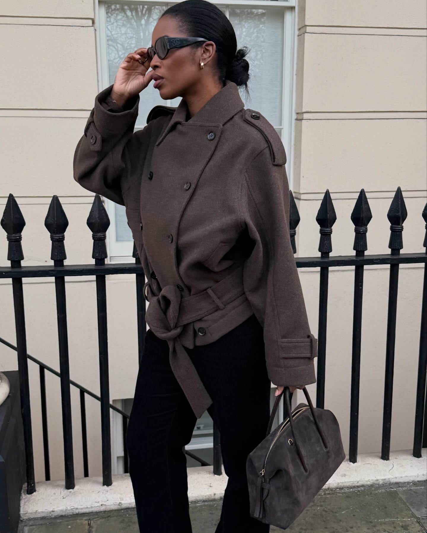 Woman wears a wool trench coat, black tailored trousers and carries suede bag
