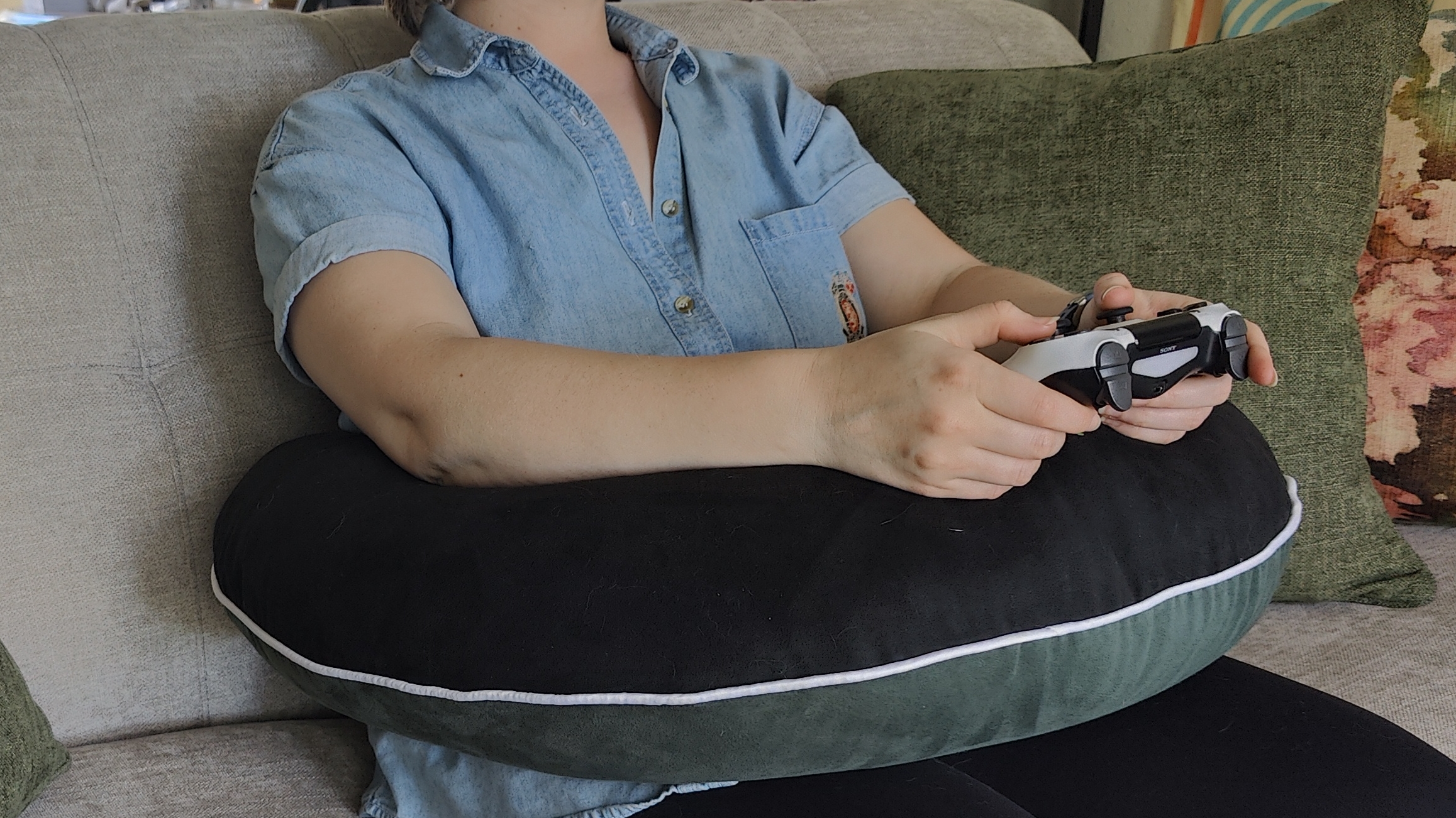 A person sitting on a couch with the Valari Gaming Pillow around their waste. Their elbows and arms are supported by the pillow and they're holding a PlayStation controller.