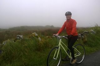 Malcom in this image for how to learn to ride a bike in a red jacket on a green bike with a stone wall and grass behind him