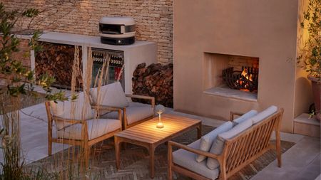 An outdoor seating area that has two garden sofas facing each other with a coffee table in the center with a lit portable lamp on top, next to a lit fireplace by an outdoor kitchen with a pizza oven
