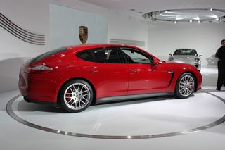 Red Porsche Panamera GTS. parked inside a silver line circle, white floor, white interior car showroom backdrop, silver parked car in the distance, circle of ceiling lights, two people in conversation