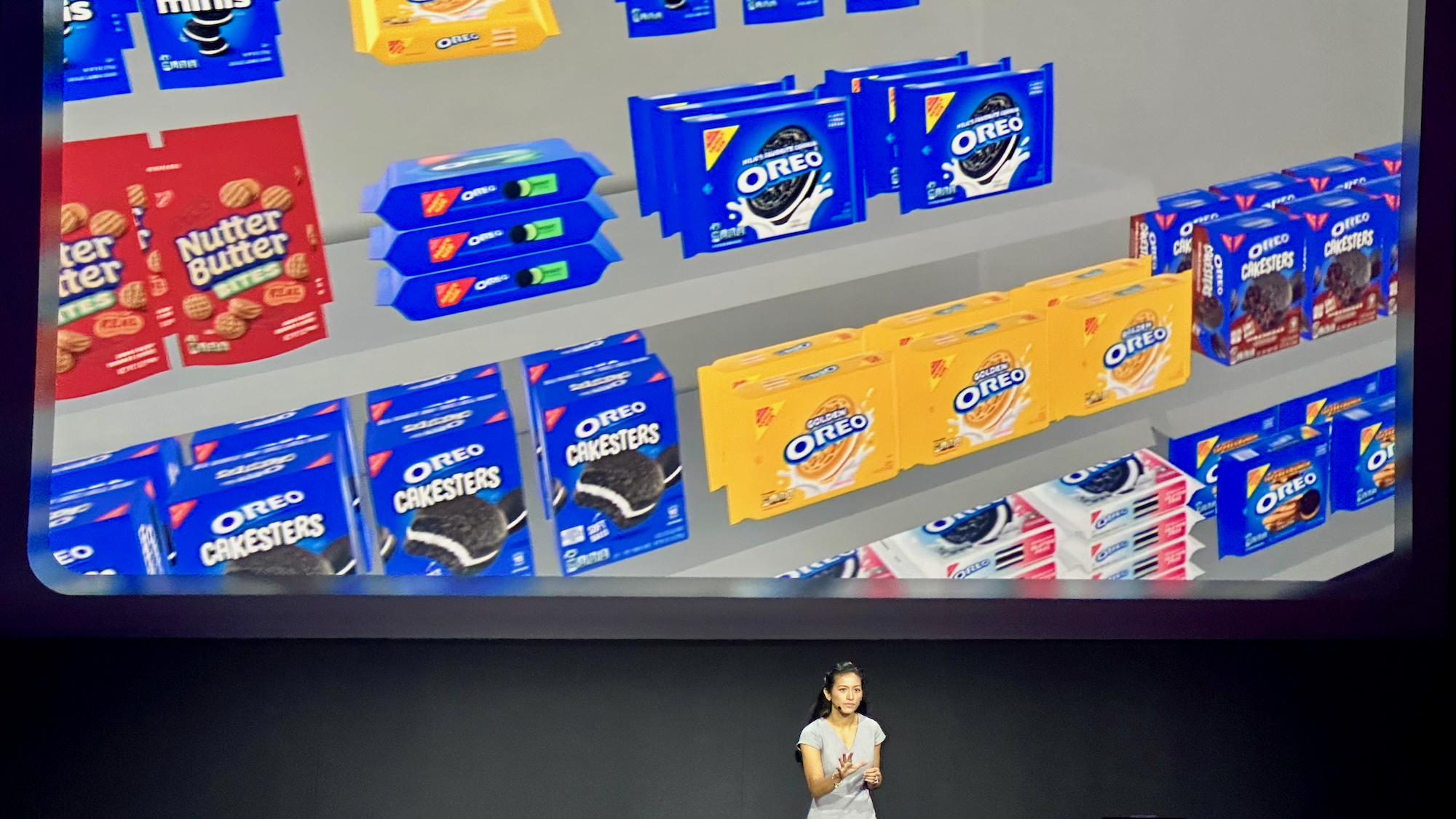 A photo of a woman on stage with a virtual storefront on a screen behind her, showing various Nabisco cookies like oreos in a sample store layout via the ShapesXR app.