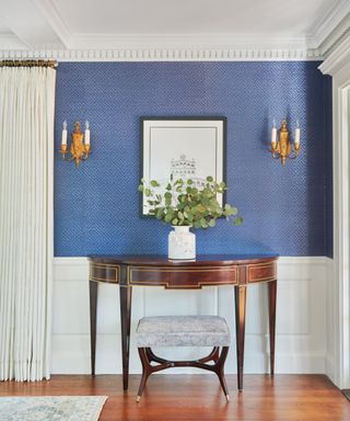 Entryway with white paneling on the lower third of the walls, and the rest covered in blue wallpaper. A vintage console table sits below a framed artwork, and a vase of eucalyptus sits on the table.