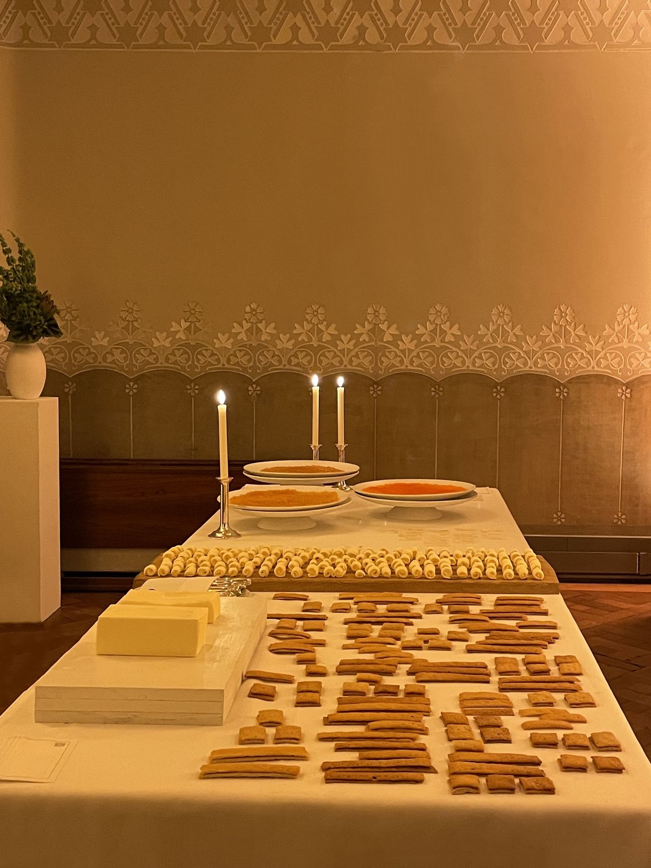 A food banquet features a table covered in a white, linen table cloth, lots of crackers presented in a choreographed way, two plates filled with food, blocks of cheese, and three lit candles in a warm setting.