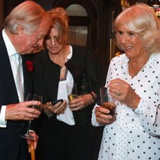 Queen Camilla wearing a white polka dot dress holding a drink and laughing next to her daughter Laura Lopes and ex-husband Andrew Parker Bowles