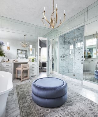 A bathroom clad with mirrored walls and white and grey marble, featuring pale blue velvet ottoman and pale pink chair at fitted white dressing table