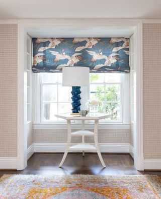 small white table in bay window with blue lampbase and blue roman blind with flamingo print and oriental rug on wooden floor
