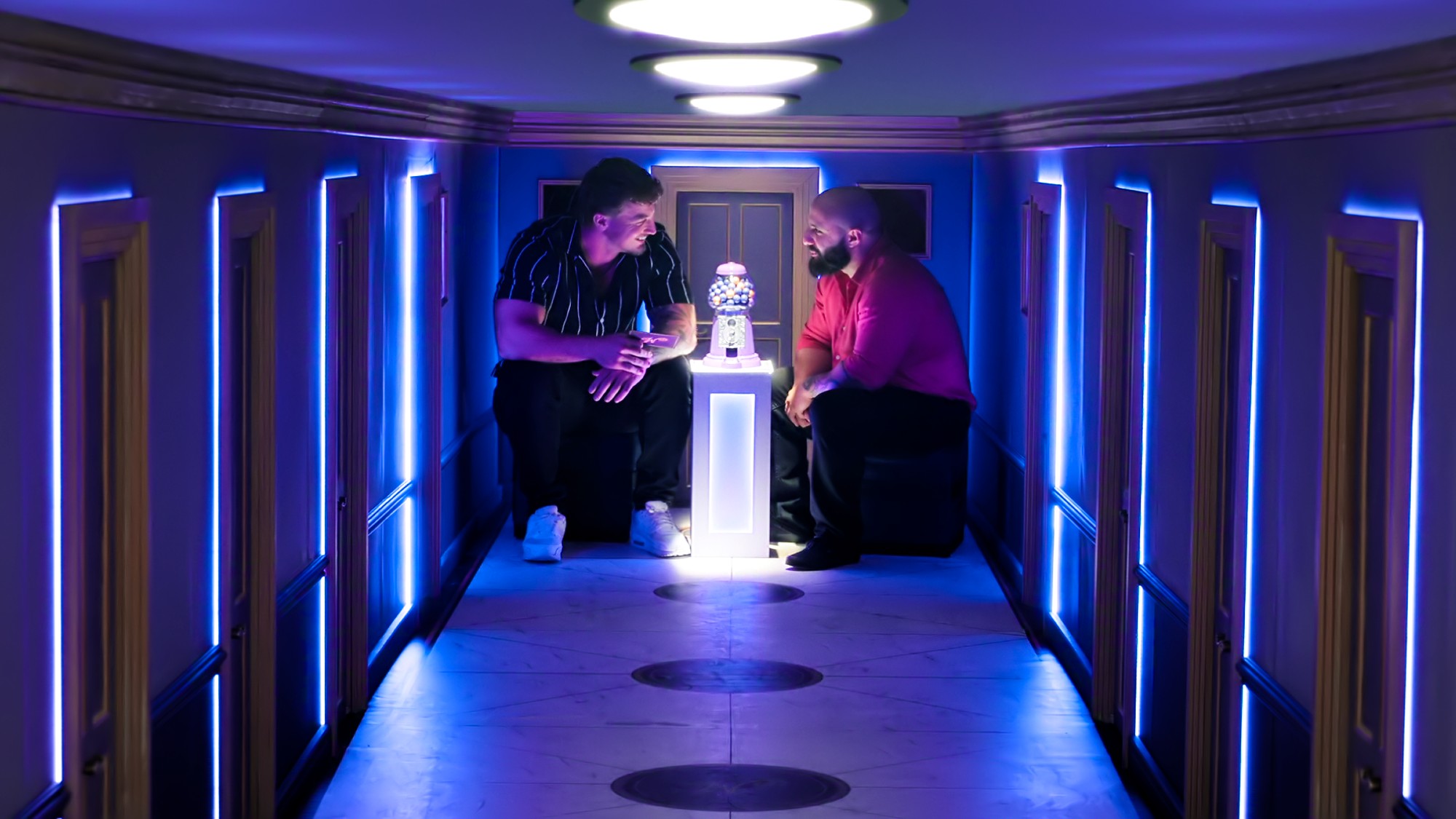 Two contestants huddle in a purple hallway that's too small for their bodies and a gumball machine sits between them.