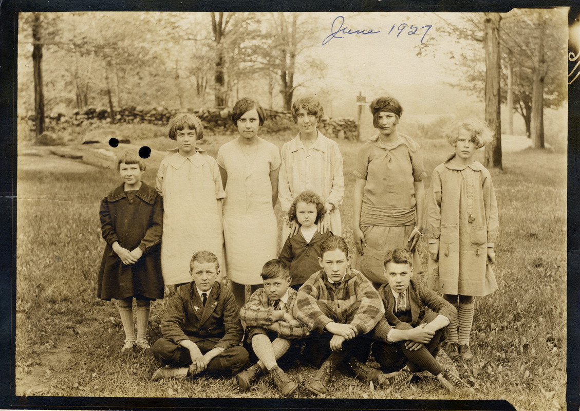 A faded sepia photograph of nine children and a young teacher posing on a grassy lawn in front of a stone wall and trees, with four boys sitting cross-legged at the front, the girls standing behind.