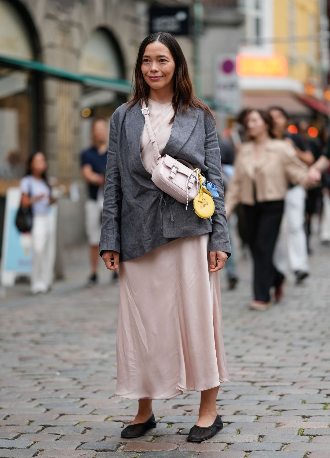 A guest wears a dark gray blazer with a subtle texture, featuring a relaxed fit and long sleeves. Underneath, a light pink satin dress with a smooth, flowing texture extends to mid-calf. A light pink crossbody bag with a structured shape and silver buckle details is worn across the body. Attached to the bag are various accessories, including a round yellow pouch from Jacquemus and a small blue Crocs shoes item. Black flat shoes complete the ensemble. Hair is styled straight and loose, outside Nicklas Skovgaard, during Copenhagen Fashion Week, on August 07, 202