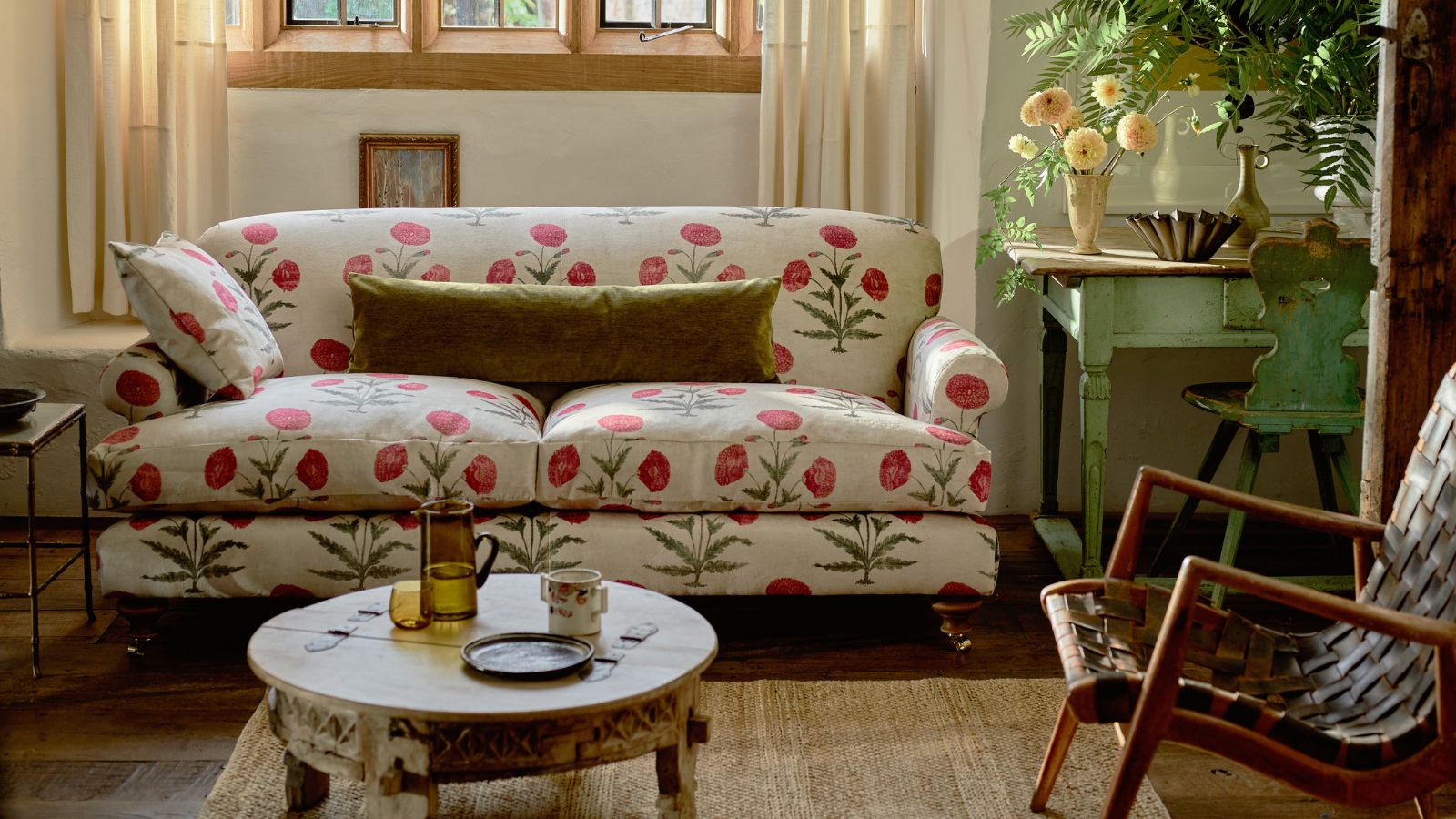 A living room with a white floral sofa with red flowers, next to a green wooden table with vases of flowers. In front of the sofa is a rug with a wooden circular coffee table, and to the right is a wooden armchair.