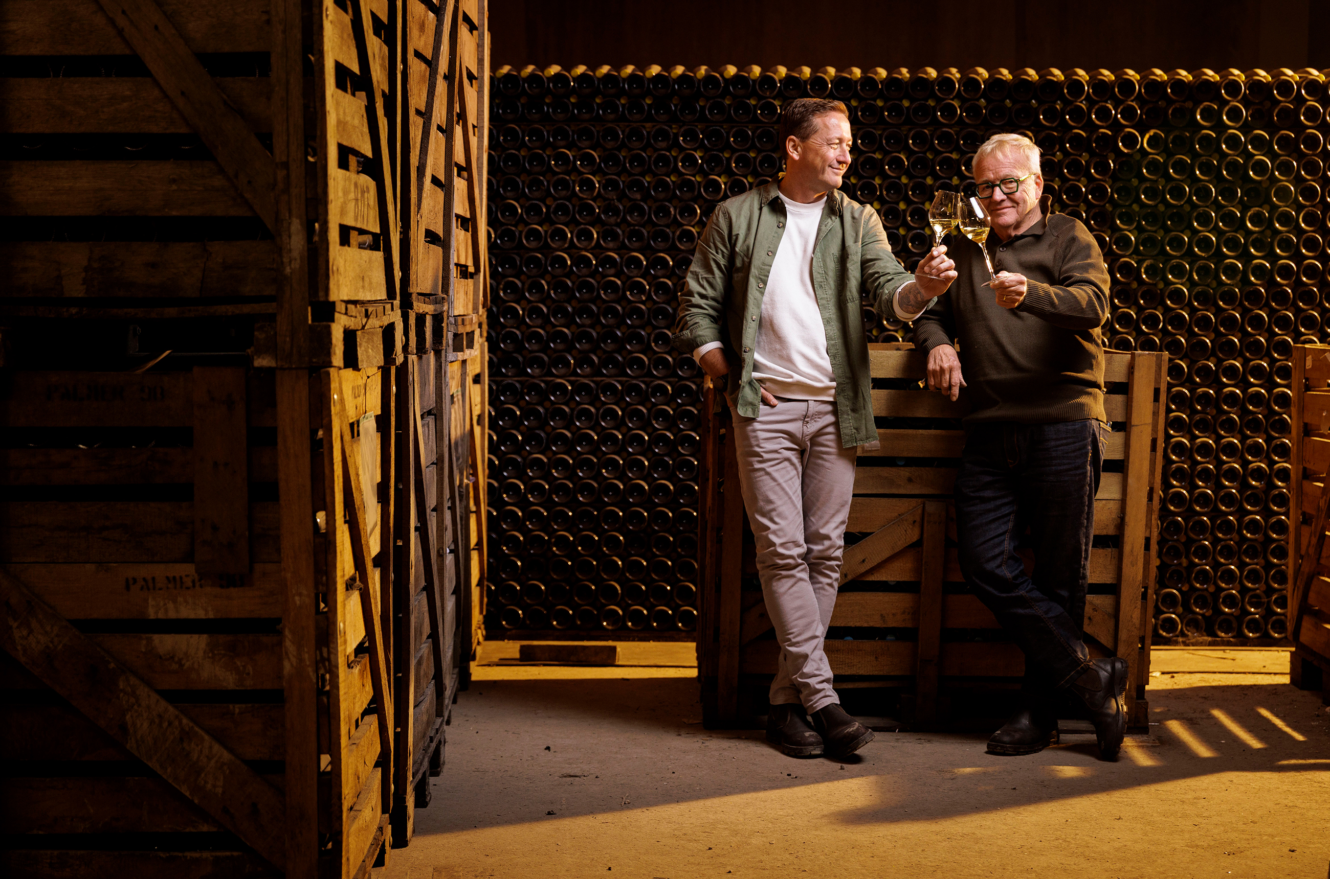 Two men holding glasses of sparkling wine in a cellar room surrounded by bottles stacked horizontally on shelves