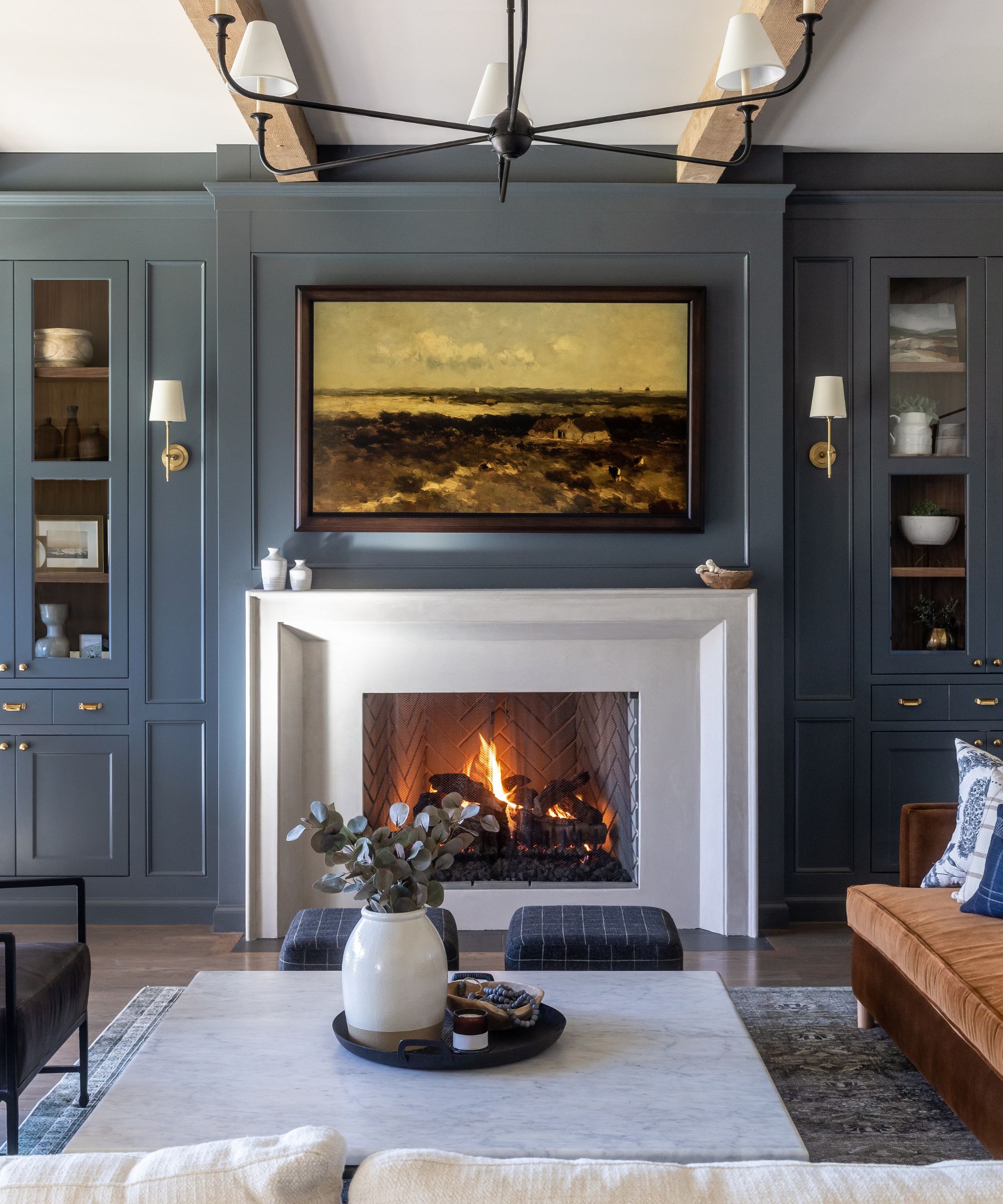 navy blue living room with a white stone fireplace and custom alcove cabinetry with a large farmhouse chandelier, wall sconces, a rug, and small blue plaid ottomans