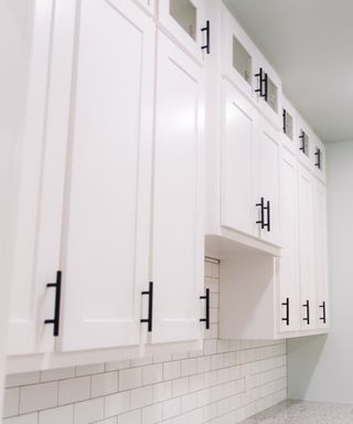 An image of a very tidy kitchen with white cabinet doors, black handles and a tiled backsplash. The doors are closed and the counter bare.
