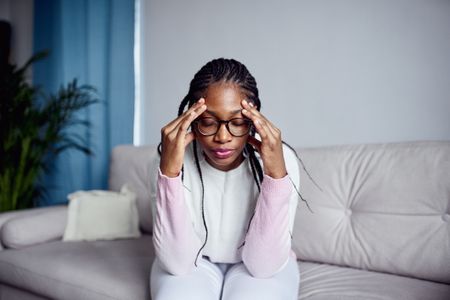 A young girl experiencing anxiety symptoms sitting on the couch at home with her head in her hands.