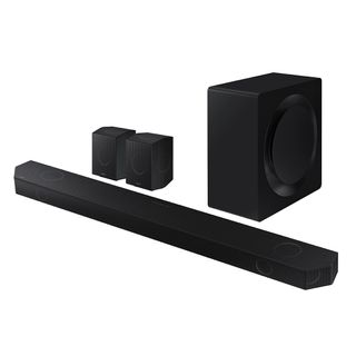 Samsung HW-Q990D soundbar system. At the front is the long soundbar, with two square satellite speakers and a boxy subwoofer behind it. All of them are completely black and the background is white.