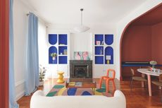 A modern living space with bright colours and a colour drenched area to the right, where the dining area is. There are floor to ceiling curtains on the left hand wall and a cream sofa facing the fireplace. 