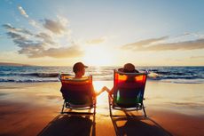 A couple sits in chair on a beach at sunset.