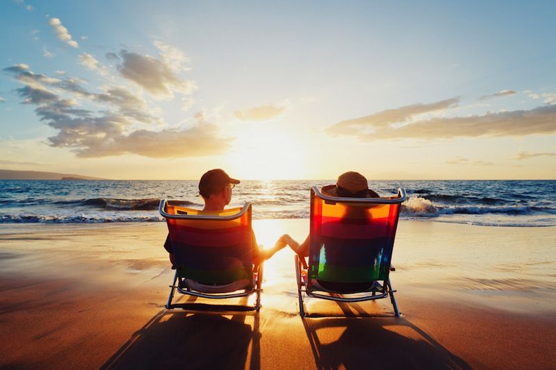 A couple sits in chair on a beach at sunset.