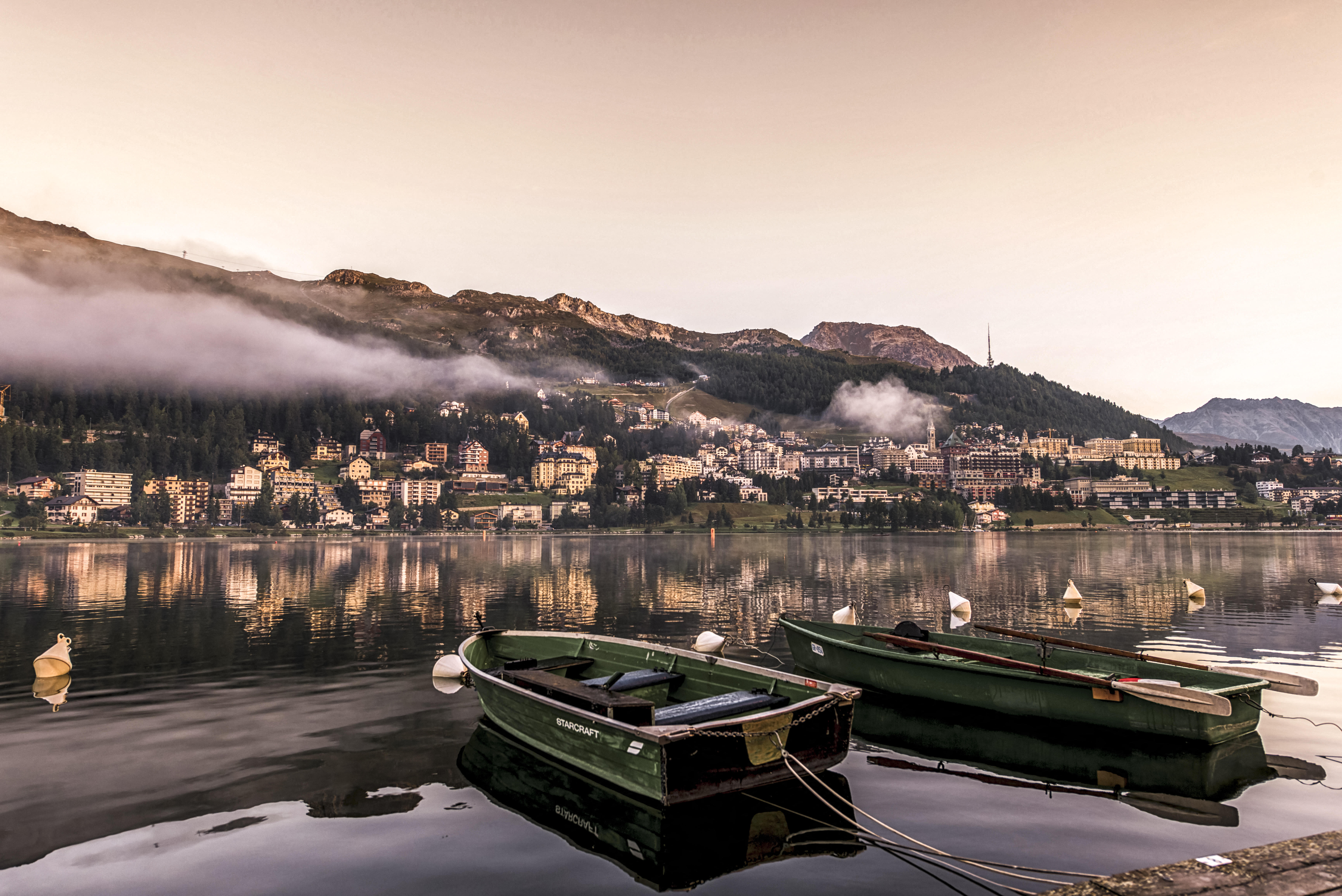 Sunrise over an alpine town and lake. There are two green rowing boats bobbing in the foreground