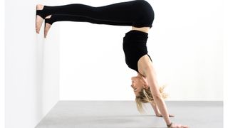 Woman facing a wall on white background performing a handstand with legs straight against wall